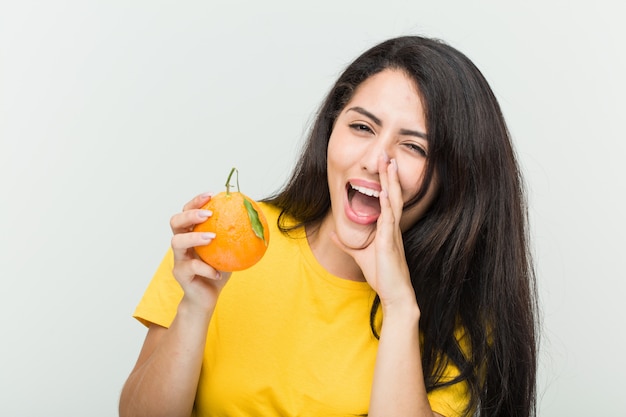 Young hispanic woman holding an orange shouting excited to front.