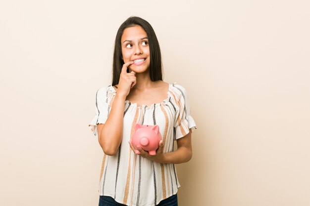 Young hispanic woman holding a piggy bank relaxed thinking about something looking at a copy space.