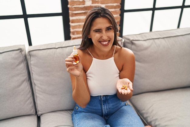 Young hispanic woman holding pills and cbd oil winking looking at the camera with sexy expression, cheerful and happy face.