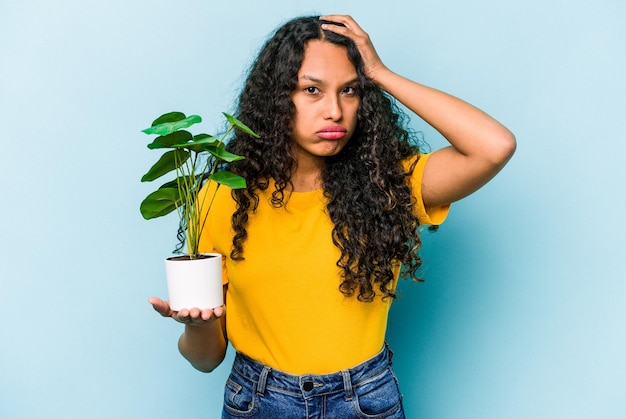 ショックを受けている青い背景に分離された植物を保持している若いヒスパニック系女性彼女は重要な会議を思い出しました