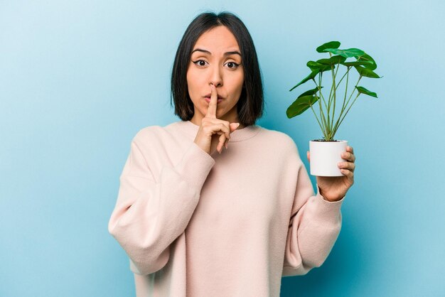 Young hispanic woman holding a plant isolated on blue background keeping a secret or asking for silence