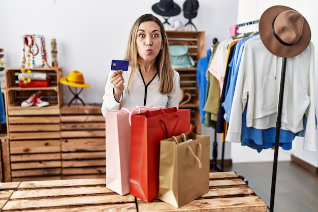 Young hispanic woman holding shopping bags and credit card at clothing store puffing cheeks with funny face. mouth inflated with air, crazy expression.