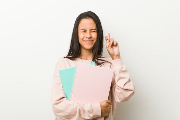 Young hispanic woman holding some notebooks crossing fingers for having luck