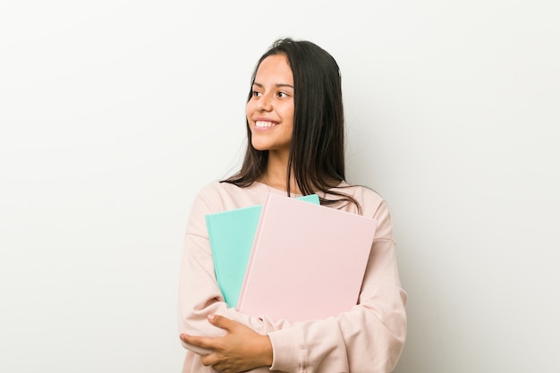 Young hispanic woman holding some notebooks smiling confident with crossed arms.