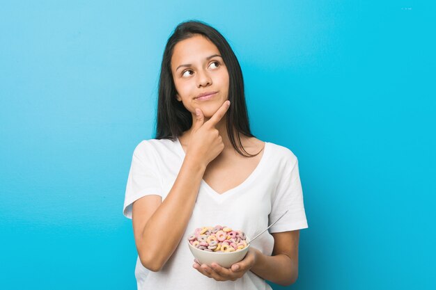Young hispanic woman holding a sugar cereal bowl looking sideways with doubtful and skeptical expression.