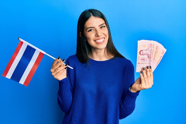 Young hispanic woman holding thailand flag and baht banknotes smiling with a happy and cool smile on face showing teeth