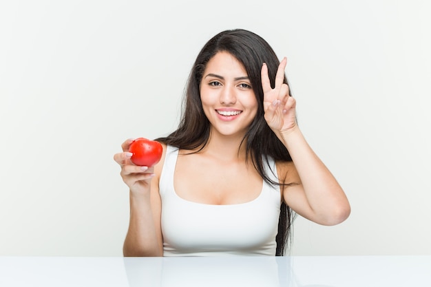 Giovane donna ispanica che tiene un pomodoro che mostra il segno di vittoria e che sorride ampiamente.