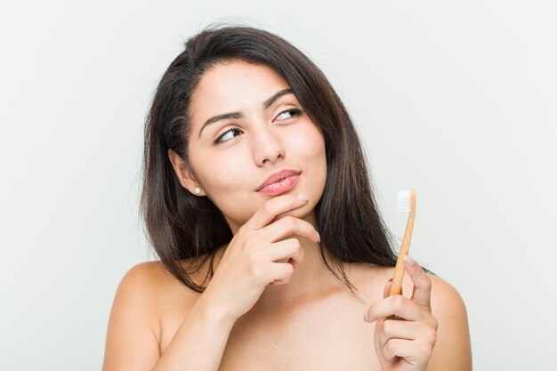 Young hispanic woman holding a toothbrush looking sideways with doubtful and skeptical expression.