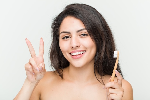 Young hispanic woman holding a toothbrush showing victory sign and smiling broadly.