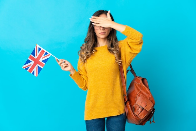 Young hispanic woman holding an United Kingdom flag on blue covering eyes by hands. Do not want to see something