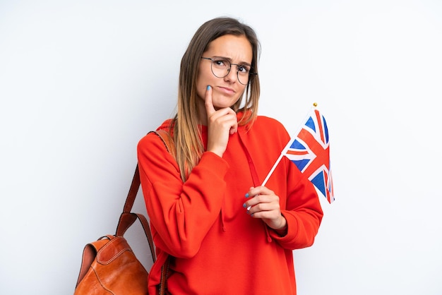 Young hispanic woman holding an United Kingdom flag isolated on white background having doubts while looking up