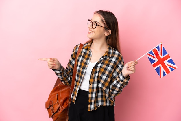 Молодая латиноамериканская женщина держит флаг Соединенного Королевства на розовом, указывая пальцем в сторону и представляет продукт