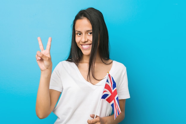 Young hispanic woman holding a united kingdom flag showing number two with fingers.