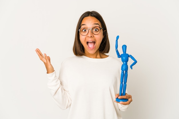 Young hispanic woman holding a wood doll surprised and shocked