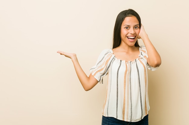 Young hispanic woman holds copy space on a palm, keep hand over cheek. Amazed and delighted.