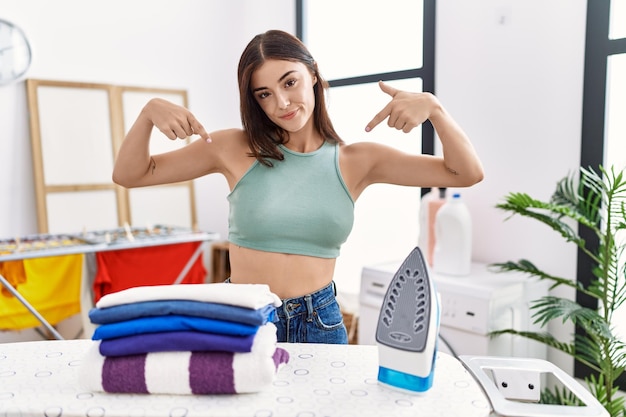 Young hispanic woman ironing clothes at laundry room looking confident with smile on face pointing oneself with fingers proud and happy