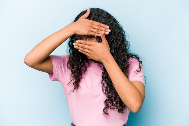 Young hispanic woman isolated on blue background blink at the camera through fingers embarrassed covering face