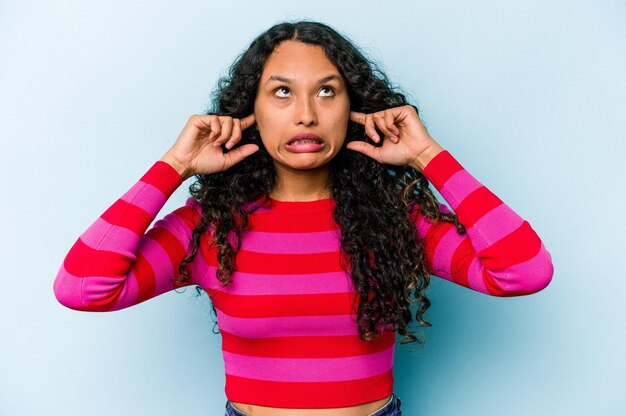 Young hispanic woman isolated on blue background covering ears with fingers stressed and desperate by a loudly ambient