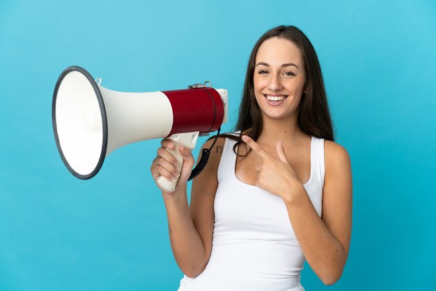 Young hispanic woman over isolated blue background holding a megaphone and pointing side