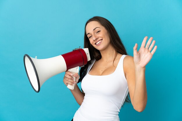 Young hispanic woman over isolated blue background holding a megaphone and saluting with hand with happy expression