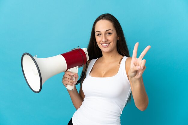 Young hispanic woman over isolated blue background holding a megaphone and smiling and showing victory sign