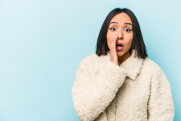 Young hispanic woman isolated on blue background is saying a secret hot braking news and looking aside