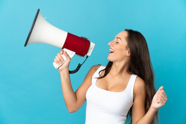 Young hispanic woman over isolated blue background shouting through a megaphone to announce something in lateral position