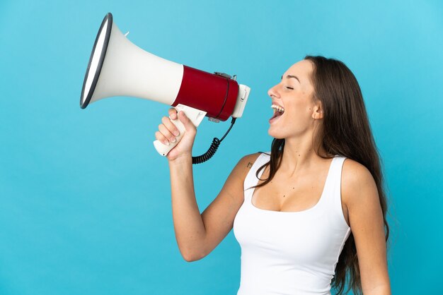 Young hispanic woman over isolated blue background shouting through a megaphone to announce something in lateral position