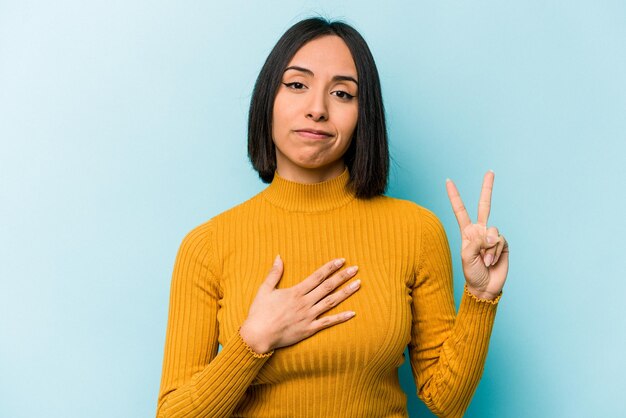 Young hispanic woman isolated on blue background taking an oath putting hand on chest