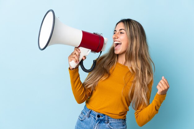 Young hispanic woman over isolated blue shouting through a megaphone to announce something in lateral position