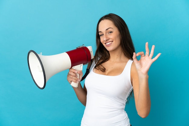 Young hispanic woman over isolated blue wall holding a megaphone and showing ok sign with fingers