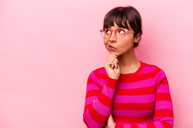 Young hispanic woman isolated on pink background looking sideways with doubtful and skeptical expression