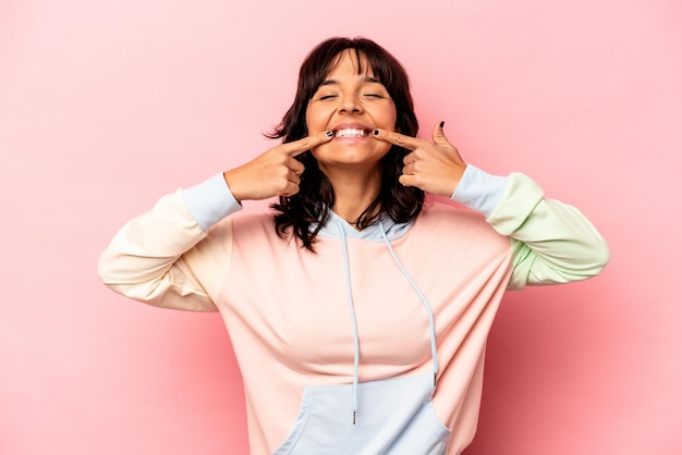 Young hispanic woman isolated on pink background smiles pointing fingers at mouth