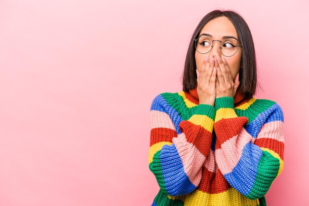 Young hispanic woman isolated on pink background thoughtful looking to a copy space covering mouth with hand