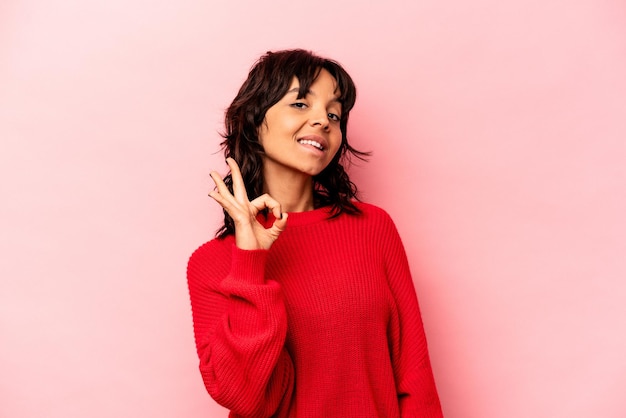 Young hispanic woman isolated on pink background winks an eye and holds an okay gesture with hand