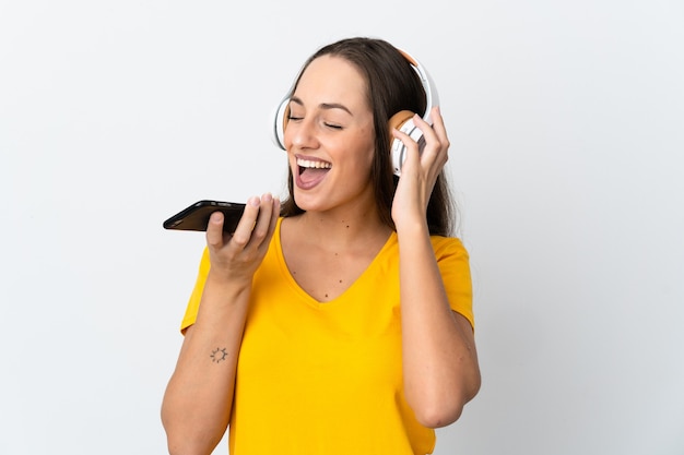 Young hispanic woman over isolated white wall listening music with a mobile and singing