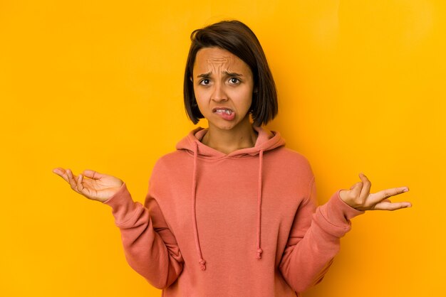 Young hispanic woman isolated on yellow confused and doubtful shrugging shoulders to hold a copy space.