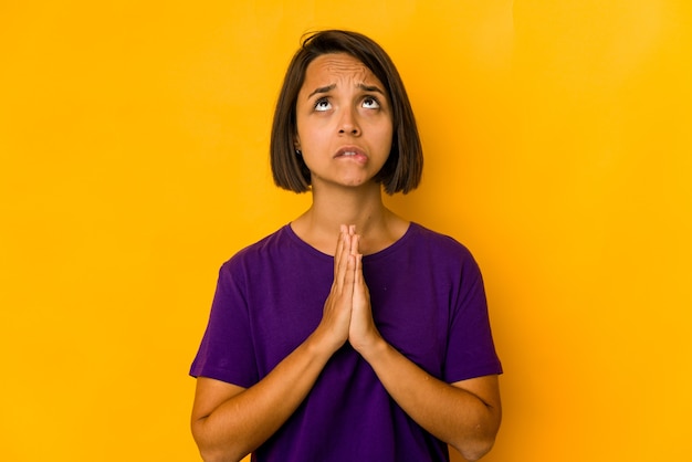 Young hispanic woman isolated on yellow holding hands in pray near mouth, feels confident.