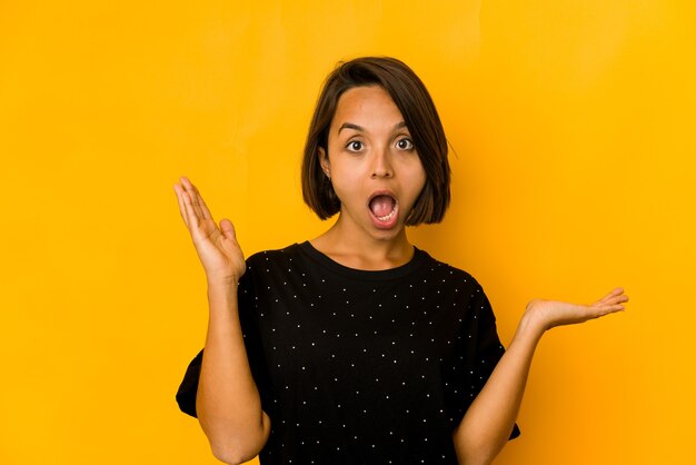 Young hispanic woman isolated on yellow holds copy space on a palm, keep hand over cheek. amazed and delighted