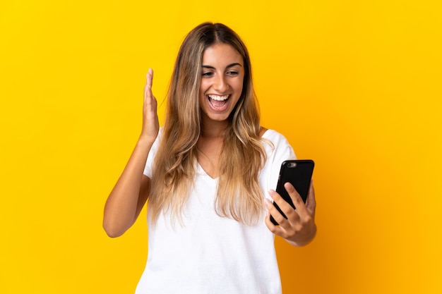 Young hispanic woman over isolated yellow looking at the camera while using the mobile with surprised expression