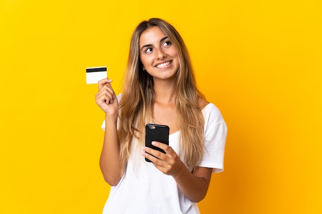 Young hispanic woman over isolated yellow wall buying with the mobile with a credit card while thinking