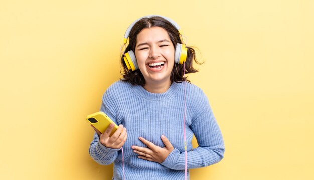 いくつかの陽気な冗談で大声で笑っている若いヒスパニック系の女性。ヘッドフォンとスマートフォンのコンセプト