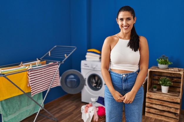 Young hispanic woman at laundry room looking positive and happy standing and smiling with a confident smile showing teeth