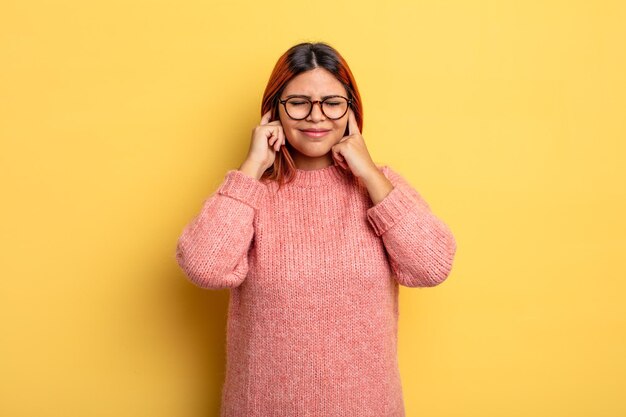 怒っているように見える若いヒスパニック系の女性は、耳をつんざくような音や大音量の音楽で両耳を覆ってストレスとイライラを感じています