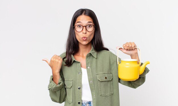 Young hispanic woman looking astonished in disbelief and holding a tea pot