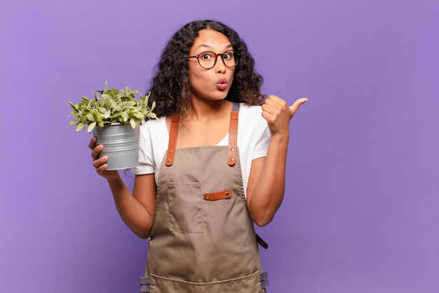 young hispanic woman looking astonished in disbelief, pointing at object on the side and saying wow, unbelievable. garden keeper concept