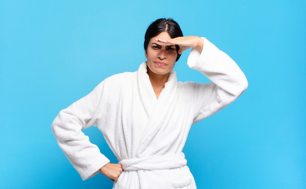 Young hispanic woman looking bewildered and astonished, with hand over forehead looking far away, watching or searching. bathrobe concept
