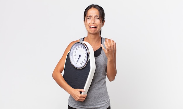 Young hispanic woman looking desperate, frustrated and stressed and holding a weight scale. diet concept