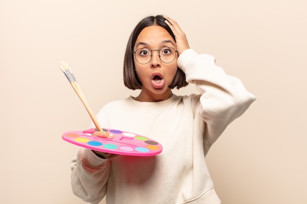 Young hispanic woman looking excited and surprised, open-mouthed with both hands on head, feeling like a lucky winner