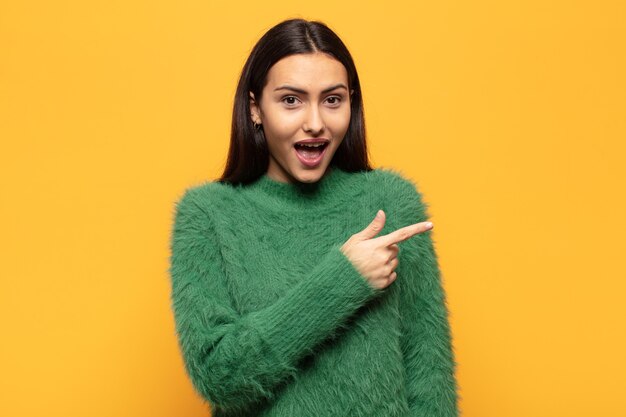 Young hispanic woman looking excited and surprised pointing to the side and upwards to copy space